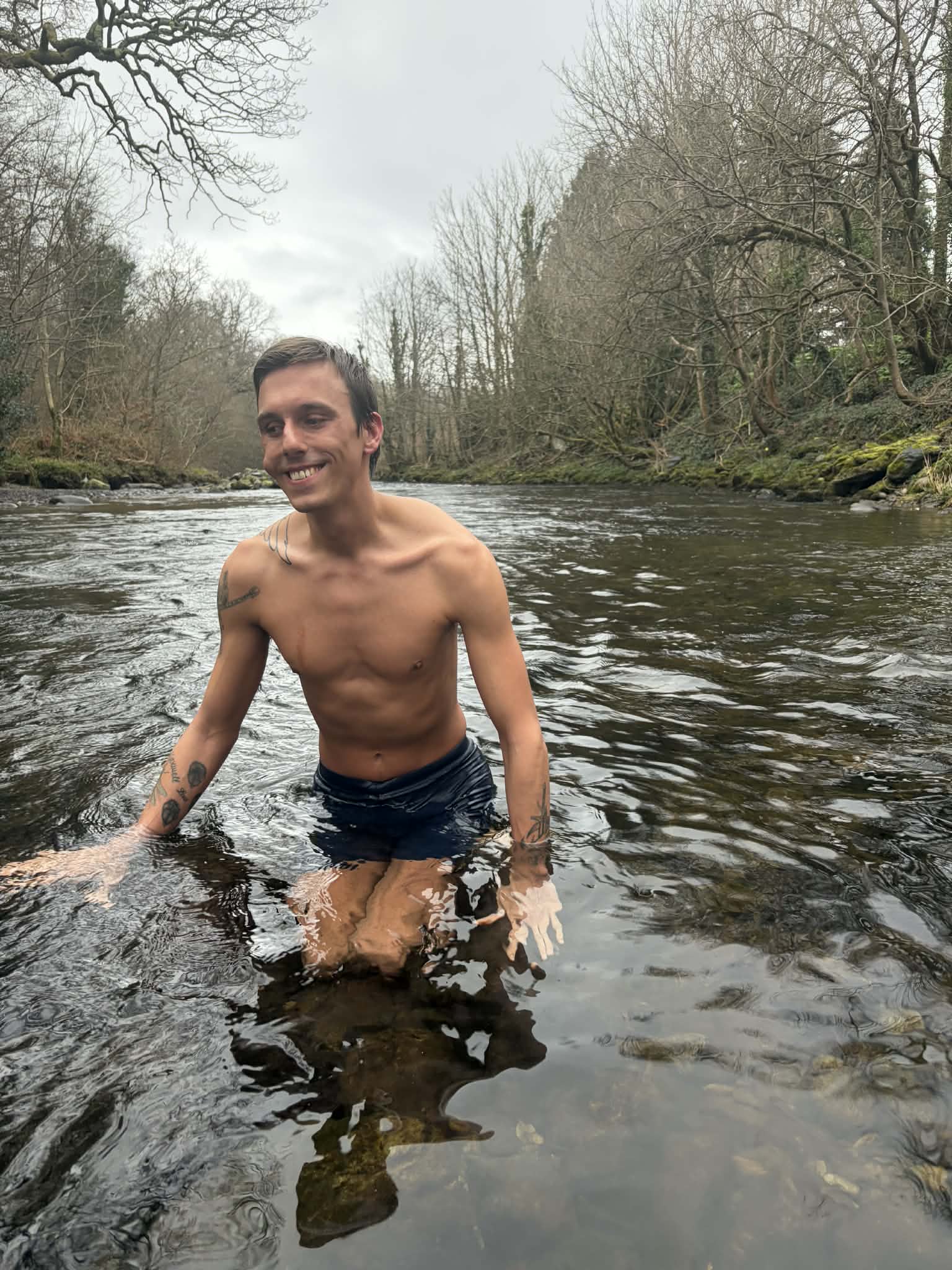 Hunter waist deep in the Afon Ogwen in winter, smiling, bare winter trees lining both banks